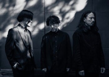 A black and white photograph of the band mouse on the keys, featuring three men dressed in dark clothing standing side by side. They are standing in front of a stone wall that is partly in the shadow and partly catches some sun rays.
