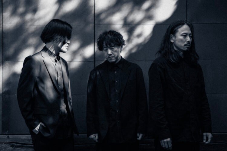 A black and white photograph of the band mouse on the keys, featuring three men dressed in dark clothing standing side by side. They are standing in front of a stone wall that is partly in the shadow and partly catches some sun rays.