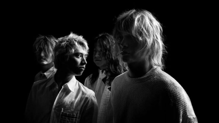 A black and white photo of the four members of ONE OK ROCK, they are standing near each other against a black background while dressed in light coloured clothing. Half of their faces are illuminated, with the other half in shadow. De vocalist of ONE OK ROCK, Taka, looks behind him, while the others are looking forward.