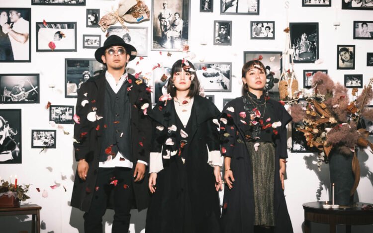 The photo features the three band members of Seven Oops, all dressed in black and standing in front of a wall adorned with monochrome photos. The picture was taken at a moment when flower petals were thrown into the air.