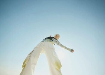 Photo of Nao Yoshioka, dressed in light-coloured clothing. The photo is taken from a low angle, with the camera pointing upwards. This perspective makes the person appear taller and more striking. Nao Yoshioka is waving her arms and looking directly into the lens.