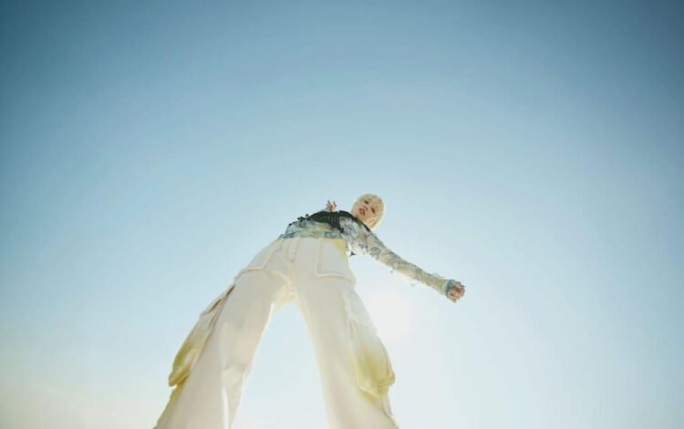 Photo of Nao Yoshioka, dressed in light-coloured clothing. The photo is taken from a low angle, with the camera pointing upwards. This perspective makes the person appear taller and more striking. Nao Yoshioka is waving her arms and looking directly into the lens.