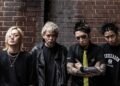 The photo features the four members of the Japanese band SiM (Silence iz Mine), all dressed in black attire and posing in front of a red brick wall.
