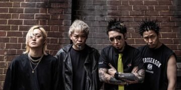 The photo features the four members of the Japanese band SiM (Silence iz Mine), all dressed in black attire and with serious facial expressions, posing in front of a red brick wall.