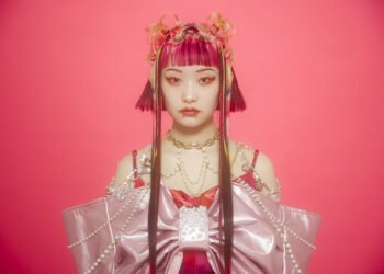 Photo of Wednesday Campanella's vocalist and frontwoman Utaha standing in front of a reddish pink background. She is dressed in an outfit that consists of the same reddish pink, but also darker and lighter pink colours. Het outfit is adorned with white, gold and yellow chains and accessories. The colours are also matched in the colours in her hair, which is styled as a bob, and face make-up.