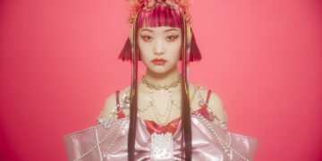 Photo of Wednesday Campanella's vocalist and frontwoman Utaha standing in front of a reddish pink background. She is dressed in an outfit that consists of the same reddish pink, but also darker and lighter pink colours. Het outfit is adorned with white, gold and yellow chains and accessories. The colours are also matched in the colours in her hair, which is styled as a bob, and face make-up.