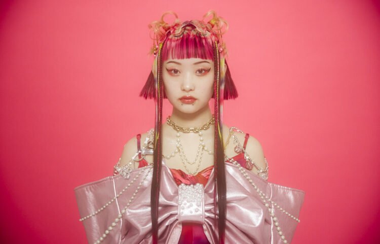 Photo of Wednesday Campanella's vocalist and frontwoman Utaha standing in front of a reddish pink background. She is dressed in an outfit that consists of the same reddish pink, but also darker and lighter pink colours. Het outfit is adorned with white, gold and yellow chains and accessories. The colours are also matched in the colours in her hair, which is styled as a bob, and face make-up.