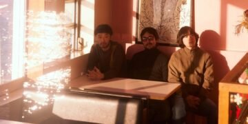 Band photo of the three-piece band Maya Ongaku sitting next to each other at a cosy cafe or diner. The seating arrangement is near a couple of windows allowing natural sunlight to shine inside the space, giving it a warm atmosphere.