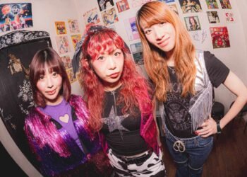 The photo features three women of THE LET'S GO's at GROOVE STOCK, probably backstage, all dressed stylishly, posing together. The wall behind them is plastered with colourful posters and drawings. Photography by Jun Sasaki (Tonewiz)