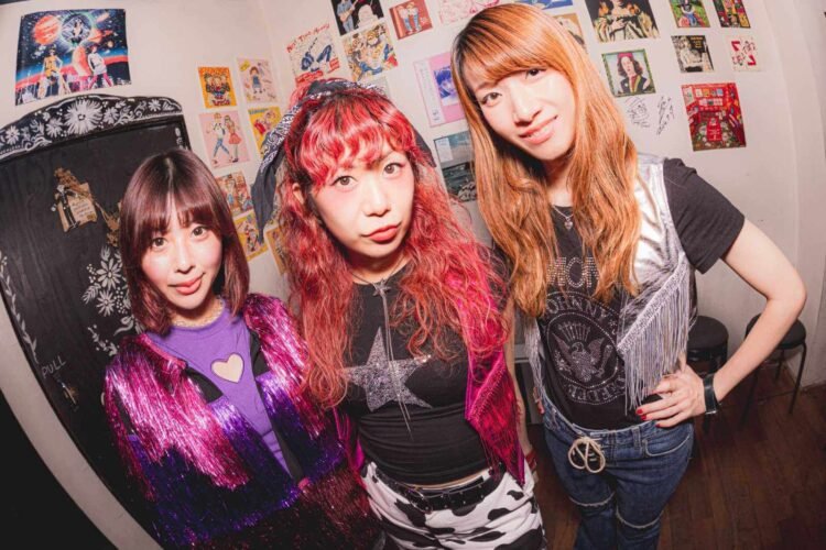 The photo features three women of THE LET'S GO's at GROOVE STOCK, probably backstage, all dressed stylishly, posing together. The wall behind them is plastered with colourful posters and drawings. Photography by Jun Sasaki (Tonewiz)