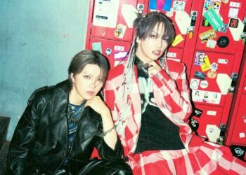 This photo features two musicians, Shou (Verde/, Alice Nine) wearing a black leather jacket and Takeru (ex-SUG) dreassed in an outfit with red and white stripes and a black shirt. They are sitting in front of red lockers that are covered with various stickers. © Takuma Hongo/Ryota Nakano