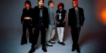 Band photo featuring the five members of Crossfaith posing against a light gray background with orange lighting on some places. From left to right: drummer Tatsuya Amano, vocalist Kenta Koie, guitarist Kazuki Takemura, guitarist Daiki Koide, toetsenist and programmer Terufumi Tamano. The band members are predominantly dressed in black outfits.