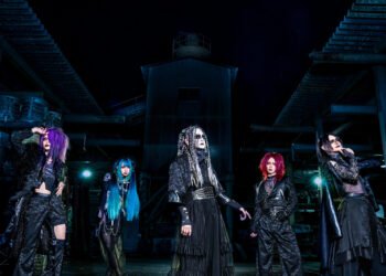 Photo of the visual kei band DAMNED, the five members are predominantly drssed in black clothing with dramatic makeup, standing in what looks like an abandonded industrial area.