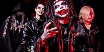 The photo portrays the four members of visual kei metal band DEVILOOF, all dressed in black attire with red and yellow/gold-coloured accents. The outfits are a combination of (fake) leather and fur, chains, belts Japanese traditional clothing. In the forefront is the vocalist Keisuke, with striking makeup: a face painted white, panda bear eyes with red lenses, black lipstick with blood effects around his mouth and on his teeth.