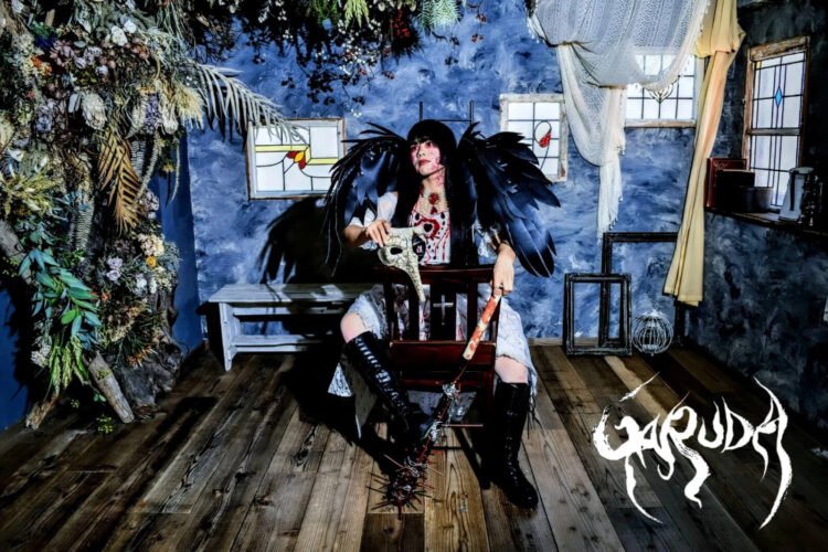 The photo shows Yuffie Sakimura as GARUDA sitting on a (prayer) chair in a room with gothic elements, plants and blue walls. She is dressed in a white dress with red elements, black wings on the back and holding a pest mask and baseball bat with spikes while looking away from the camera.