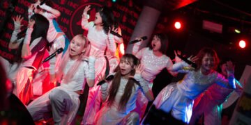 The photo depicts a moment where the idol group GANG PARADE, consisting of 11 members, are at the end of a song and posing. All members are dressed in white outfits with on the back a bright colour, it is a different colour for everyone.