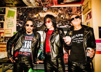 The current three men of Guitar Wolf are standing next to each other in a space that is decorated with posters: guitarist and vocalist Seiji, bassist Gotz and drummer Takuro, dressed in black leather.