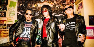 The current three men of Guitar Wolf are standing next to each other in a space that is decorated with posters: guitarist and vocalist Seiji, bassist Gotz and drummer Takuro, dressed in black leather.