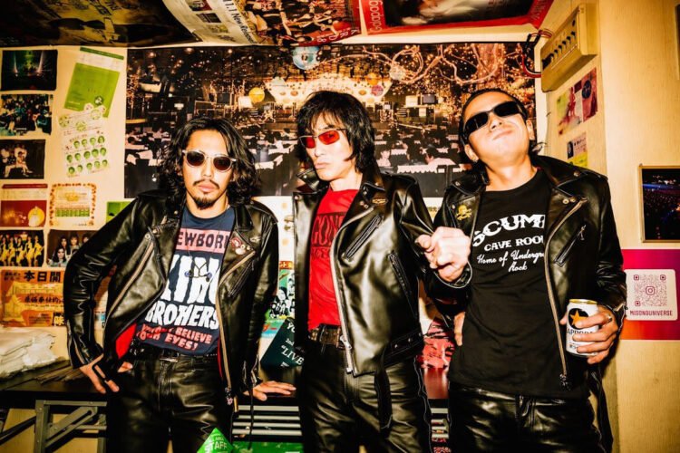 The current three men of Guitar Wolf are standing next to each other in a space that is decorated with posters: guitarist and vocalist Seiji, bassist Gotz and drummer Takuro, dressed in black leather.