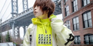 Photo of NANO standing in a street in New York on a rainy day. Behind NANO is a cable bridge and she is dressed in a white jacket over a neon yellow hoodie. Photo by Joey (@evillittlejawa)