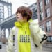 Photo of NANO standing in a street in New York on a rainy day. Behind NANO is a cable bridge and she is dressed in a white jacket over a neon yellow hoodie. Photo by Joey (@evillittlejawa)