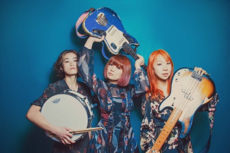 The three members of TsuShiMaMiRe standing side by side against a blue wall, each holding the musical instrument they play. The singer and guitarist Mari holds her guitar above her head. All three women are wearing colourful, patterned outfits with seemingly floral designs.