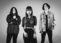 A black-and-white photo of the three band membes of GUNGIRE. They are standing in front of what seems to be a white or light backdrop. They are standing side by side, mostly dressed in dark clothing, only the vocalist is standing a bit more foreward. | Photography by Saskia (sas.silence on Instagram)