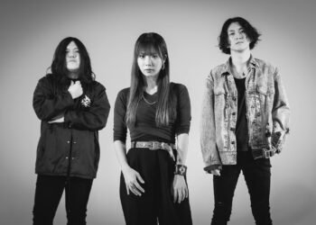 A black-and-white photo of the three band membes of GUNGIRE. They are standing in front of what seems to be a white or light backdrop. They are standing side by side, mostly dressed in dark clothing, only the vocalist is standing a bit more foreward.  | Photography by Saskia (sas.silence on Instagram)
