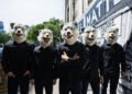 The photo shows the five members of MAN WITH A MISSION standing in a row on a street. They are dressed in dark clothing, including jeans and shirts with a MAN WITH A MISSION on it, while wearing their full-face wolf masks.