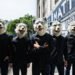 The photo shows the five members of MAN WITH A MISSION standing in a row on a street. They are dressed in dark clothing, including jeans and shirts with a MAN WITH A MISSION on it, while wearing their full-face wolf masks.