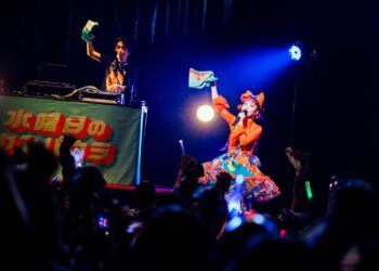 Photo of Wednesday Campanella at Asakusa Public Hall in Tokyo in April 2025. The photo captures a lively scene of a performance on a brightly lit stage. On the stage, Utaha is dressed in a colourful outfit and Kenmochi is on hte background on his DJ booth. Both are waving with a towel/muffler. Photo by Masato Yokoyama, courtesy of Tsubasa Records