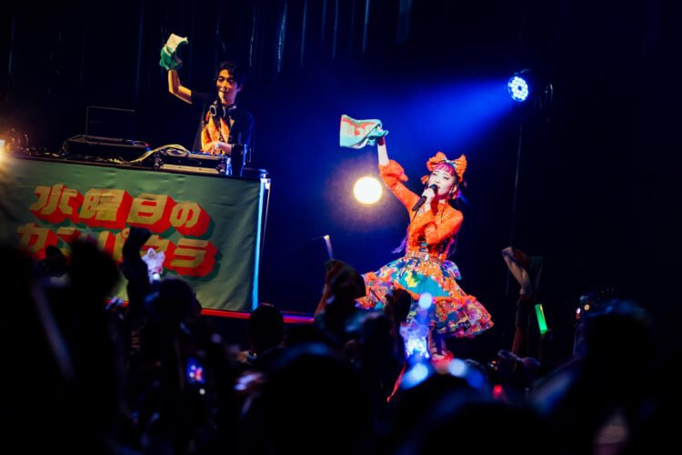 Photo of Wednesday Campanella at Asakusa Public Hall in Tokyo in April 2025. The photo captures a lively scene of a performance on a brightly lit stage. On the stage, Utaha is dressed in a colourful outfit and Kenmochi is in the background on his DJ booth. Both are waving with a towel/muffler. Photo by Masato Yokoyama, courtesy of Tsubasa Records