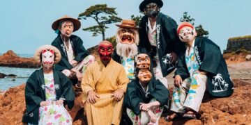 A group of people that are part of Minyo Crusaders with Hyottoko and Otafuku masks and happi coats posing for a picture at a beach with rock formations.