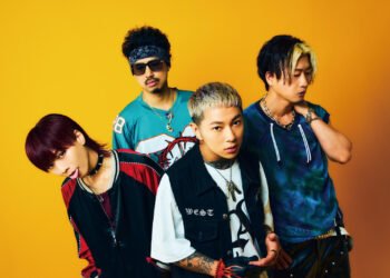 Band photo of ASH DA HERO featuring four members standing together against a vibrant yellow/orange background. They each wear different shirts in different colours, which reflect their characters.