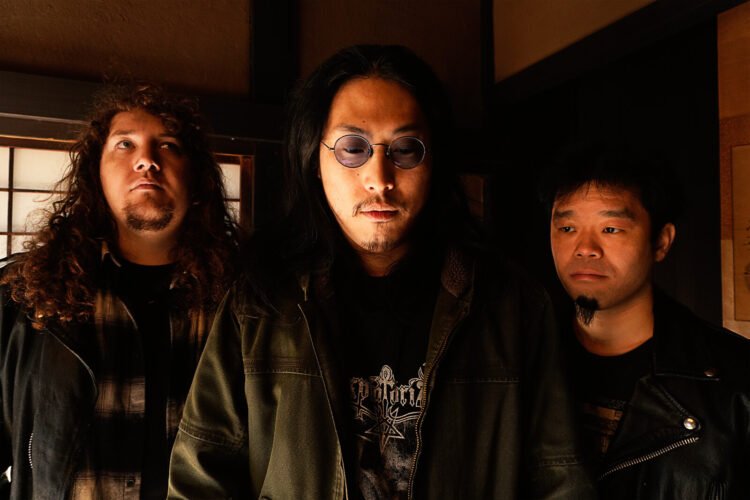 The photo features the three men of the band Hebi Katana standing in a Japanese traditional room in natural lighting. They are standing close together and are all wearing dark-coloured clothing. Photo by Hiromi Furukawa
