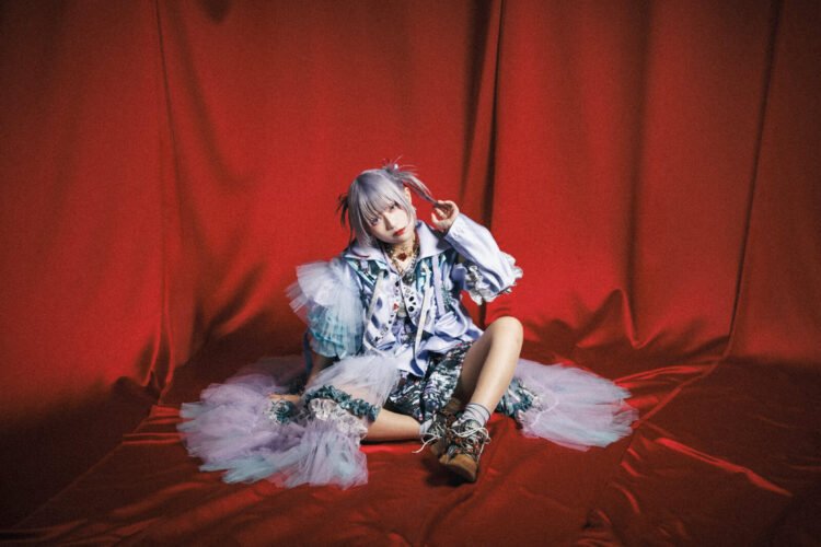 The photo features MAD JAMIE's Akubi Kanjosen with white-blond hair sitting on a red cloth. She is wearing an elaborate outfit with pastel colours and white, blue and black, frills, gems, chiffon, and ribbons.