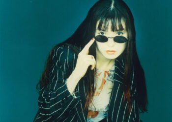 nOrikO aka POiSON GiRL FRiEND  with long dark hair, dressed in a black jacked with white stripes and wearing dark sunglasses. She is pointing her index finger to the side of her sunglasses which are lowered so that you can easily see her eyes. This photo was taken in or around 1994 and was probably used for the promotion of POiSON GiRL FRiEND's album "Love Me".