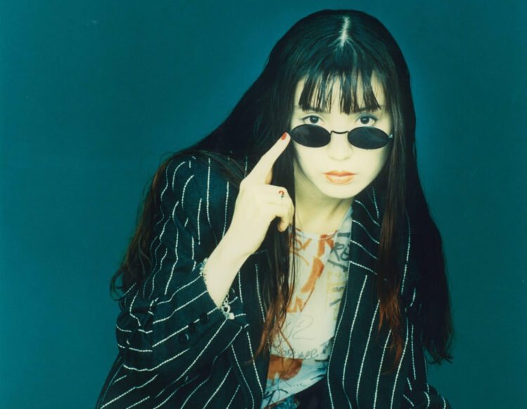 nOrikO aka POiSON GiRL FRiEND with long dark hair, dressed in a black jacket with white stripes and wearing dark sunglasses. She is pointing her index finger to the side of her sunglasses, which are lowered so that you can easily see her eyes. This photo was taken in or around 1994 and was probably used for the promotion of POiSON GiRL FRiEND's album "Love Me"
