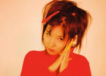 Close-up photo of Aya Gloomy. She looks directly into the camera while holding her hands together on the left side of her face and tilting her head slightly. She wears a red dress and her hair are in buns/pigtails with red hair accesoires, while her nails are in a yellow colour. Photography by Mahne Frame