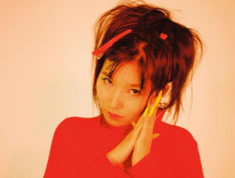 Close-up photo of Aya Gloomy. She looks directly into the camera while holding her hands together on the left side of her face and tilting her head slightly. She wears a red dress and her hair are in buns/pigtails with red hair accesoires, while her nails are in a yellow colour. Photography by Mahne Frame