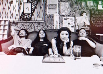 The photo shows the four members of Hibushibire casually relaxing on a sofa with a table in front of them. The photo in black-and-white with a colourshift filter showing a bit of red and green. While the three men are leaning back on the sofa, the female band member is leaning forward. On the table are an ashtray full of cigarettes and a drink.