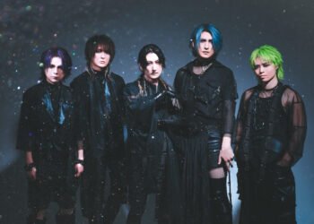 The photo shows a five members of the visual kei band Matenrou Opera. They are standing together closely, all dressed in black outfits. The background is also dark and it appears that there is a presence of bokeh.