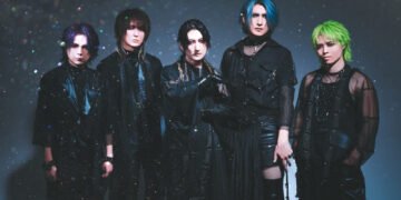 The photo shows a five members of the visual kei band Matenrou Opera. They are standing together closely, all dressed in black outfits. The background is also dark and it appears that there is a presence of bokeh.
