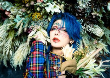 This photo features the musician Kouki of the visual kei band D=OUT with striking blue hair posing with a bouquet of flowers and also being surrounded by a floral decoration behind him. Kouki's face is adorned with artistic makeup that is inspired by flowers and he wears a military jacket with tartan patterns (colours: blue, red, yellow).