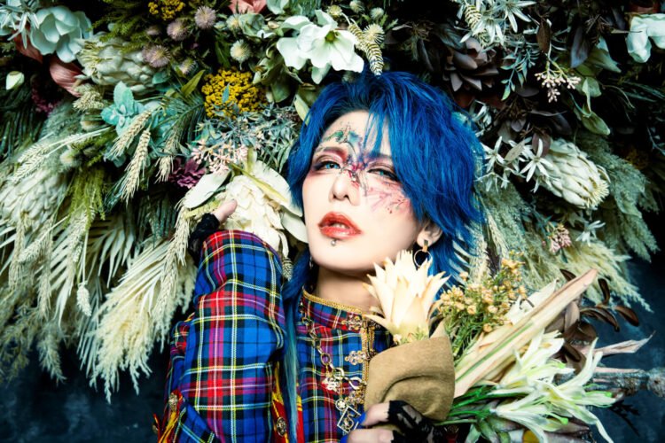 This photo features the musician Kouki of the visual kei band D=OUT with striking blue hair posing with a bouquet of flowers and also being surrounded by a floral decoration behind him. Kouki's face is adorned with artistic makeup that is inspired by flowers and he wears a military jacket with tartan patterns (colours: blue, red, yellow).