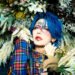 This photo features the musician Kouki of the visual kei band D=OUT with striking blue hair posing with a bouquet of flowers and also being surrounded by a floral decoration behind him. Kouki's face is adorned with artistic makeup that is inspired by flowers and he wears a military jacket with tartan patterns (colours: blue, red, yellow).