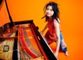 Photo of Hiromi Uehara smiling and standing behind a grand piano while also playing it. The piano is occupying the left half of the photo. The room has a bright orange floor and wall. The orange colour also comes back in the piano and the top that Hiromi Uehara is wearing, making it a bright picture.