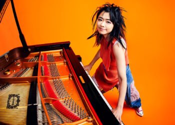 Photo of Hiromi Uehara smiling and standing behind a grand piano while also playing it. The piano is occupying the left half of the photo. The room has a bright orange floor and wall. The orange colour also comes back in the piano and the top that Hiromi Uehara is wearing, making it a bright picture.