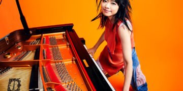 Photo of Hiromi Uehara smiling and standing behind a grand piano while also playing it. The piano is occupying the left half of the photo. The room has a bright orange floor and wall. The orange colour also comes back in the piano and the top that Hiromi Uehara is wearing, making it a bright picture.