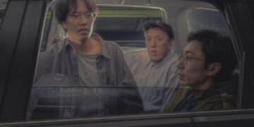 Photo of the three members of Kukangendai in a black car. The three men are dressed in casual clothing and sitting next to each other. One of them, sitting in the middle, is leaning forward and looking out of the right-hand window of the car. Another person is looking straight ahead, while the third person is looking at the person in the middle.